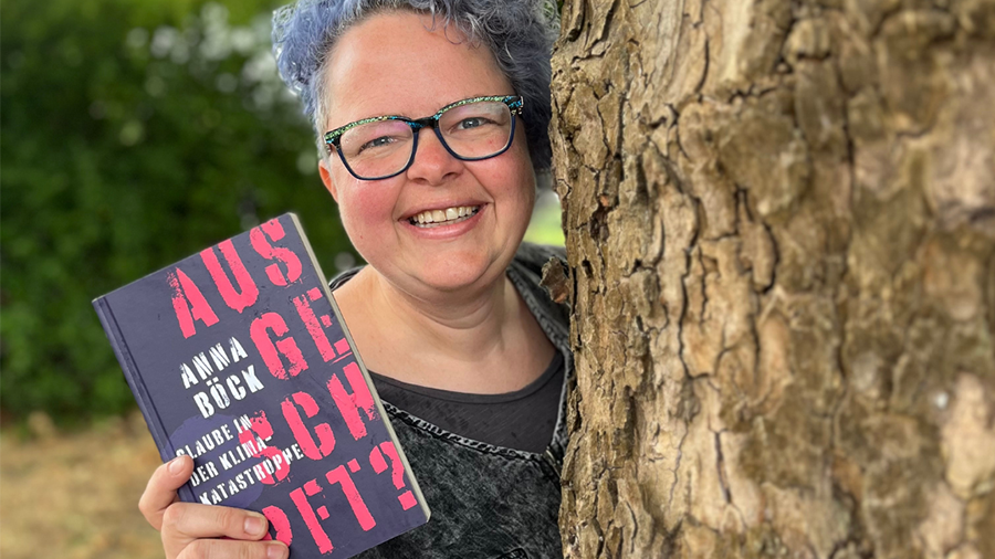 Anna Böck steht neben Baum und hält Buch "ausgeschöpft" in der Hand.